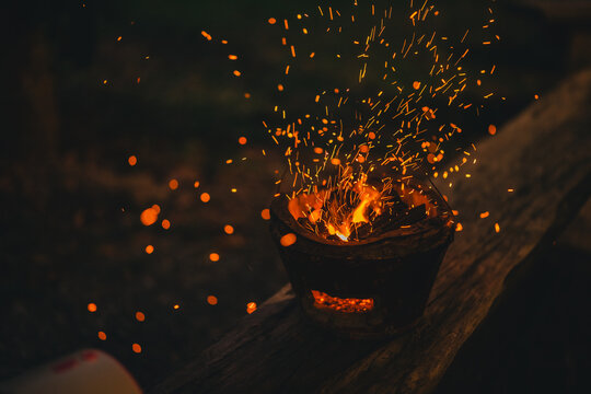Fire spark flame ember charcoal brazier heat campfire night outdoor glow from burning charcoal in metal bowl on wooden surface with flying orange spark and dark rustic mood