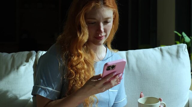 Beautiful young woman with long red hair wearing pajamas, sitting on a couch in the morning sunlight, smiling while scrolling on her smartphone and drinking a cup of coffee