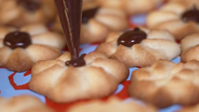 Close-up view of chocolate icing being piped onto the center of freshly baked cookies arranged on a vibrant surface, showcasing the meticulous decoration process in a kitchen setting