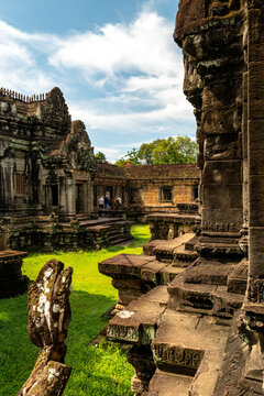 View of the ancient stone ruins of a temple with intricate carvings, mossy steps, and lush green grass under a bright blue sky in Krong Siem Reap, Siem Reap Province, Cambodia.