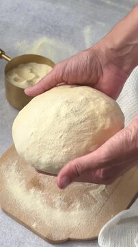 A woman's hands pick up a ball of raw dough, dusted with flour, from a wooden cutting board. Kneading dough for homemade bread or pizza.
