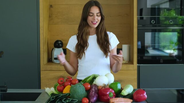 Beautiful young female blogger talking to the camera and explaining a vegan recipe while standing in her modern kitchen with fresh organic vegetables on the countertop for healthy cooking