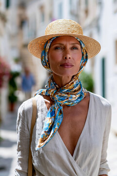 Photorealistic candid street style scene of a woman walking through a sunlit Italian coastal town wearing a classic straw canotier hat and a colorful silk headscarf tied at her nec