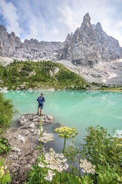 Young man hiker at the turquoise-green Sorapis lake with flowers, Lago di Sorapis, mountain peak Dito di Dio, Dolomites, Belluno, Italy