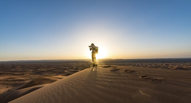 Young man standing on sand dune and photographed, sunbeams at sunrise, Erg Chebbi, Merzouga, Sahara, Morocco