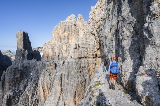 Two mountaineers on an exposed rock band in the secured Via Ferrata Bocciere Centrale via ferrata, spectacular mountain landscape with steep rocky peaks, Brenta Mountains, Parco Naturale Brenta-Adamello, Trentino, Italy