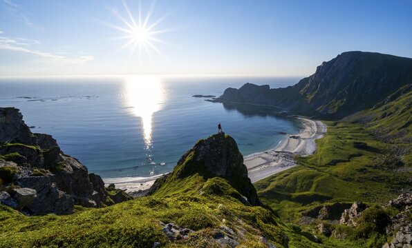 Climber on a rock, view of picturesque coastal landscape with sun star, steep coast with peak M&aring;tind or M&aring;tinden, H&oslash;yvika beach and sea, hike to the summit of M&aring;tind, And&oslash;ya island, Vester&aring;len, Nordland, Norway