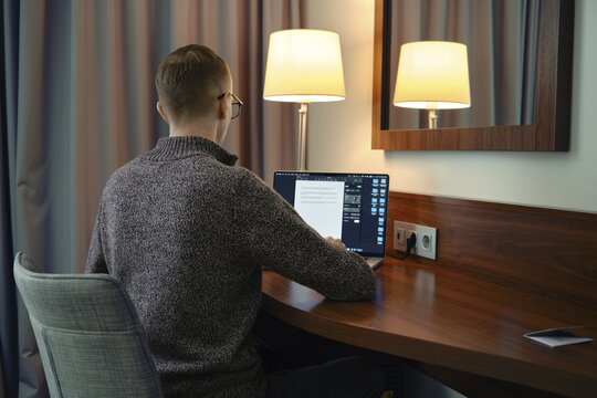 A person sits at a wooden desk in a hotel room, focused on his laptop screen. Soft lamp light creates a warm ambiance as he reviews documents late in the evening