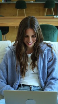 Smiling young woman with long hair sitting comfortably on the sofa while typing on her laptop keyboard, working or studying online in a cozy and modern living room environment at home