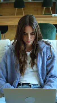 Beautiful smiling woman typing on a laptop keyboard while sitting on a comfortable sofa in her living room, enjoying a modern digital nomad lifestyle and remote work flexibility