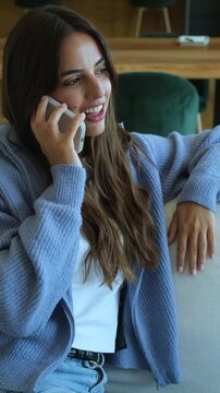 Cheerful young woman with long hair sitting on the sofa in her living room, enjoying a pleasant conversation on her smartphone and smiling with genuine happiness and contentment