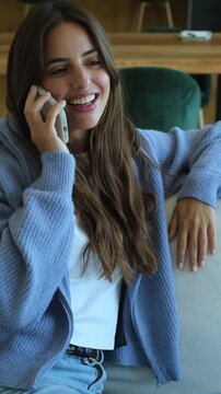 Beautiful young woman with long hair sitting comfortably on the sofa in her living room, enjoying a cheerful and happy conversation while talking on her mobile phone and smiling