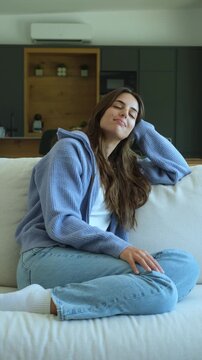 Serene young woman sitting on a comfortable sofa in her modern living room, daydreaming and enjoying a quiet moment of peace and tranquility at home with a gentle, happy smile on her face
