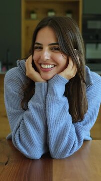 Beautiful young woman with long brown hair smiling and looking at the camera, resting her head on her hands while sitting at a wooden table in a modern kitchen, expressing happiness and serenity
