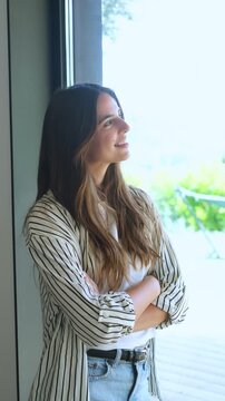 Portrait of a beautiful young woman with her arms crossed, smiling as she looks out the window, feeling relaxed, happy, and confident while enjoying a peaceful moment at home or in her office