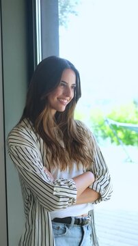 Serene and confident young woman with her arms crossed, smiling as she gazes thoughtfully out a large window, contemplating future opportunities and enjoying a peaceful moment