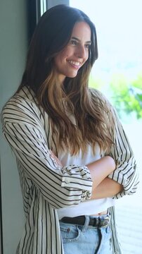 Beautiful young woman standing with arms crossed, smiling confidently while looking out a bright window, enjoying a moment of peaceful reflection and contentment in a modern setting