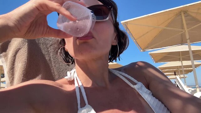 Close up of young woman sunbathing on sun lounger and drinking cold water from plastic glass to prevent dehydration during hot summer day on beach