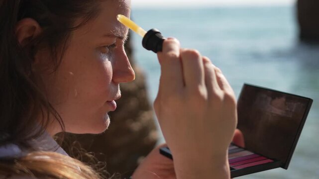 Skincare serum beach young woman applying facial oil with a dropper while sitting on a rocky coast for glowing summer skin