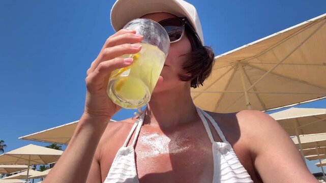 Young woman in white bikini and cap sunbathing on sun lounger and drinking cold refreshing lemonade from glass to cool down and beat summer heat on beach resort