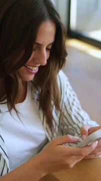 Beautiful young woman with long brown hair smiling while using her mobile phone, scrolling and typing a message on the touchscreen while sitting at a wooden table in a brightly lit cafe
