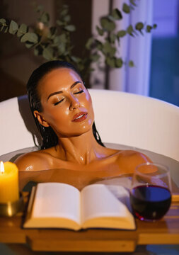Beautiful woman relaxes in a freestanding bathtub with her eyes closed, soaking in a calm ambience. A wooden tray holds an open book, a glowing yellow candle, and a glass of red wine