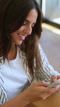 Beautiful happy woman sitting at a table using her mobile phone, scrolling and typing messages. Millennial girl enjoys chatting on social media apps, smiling while reading content online