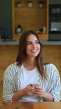 Joyful woman sitting at a wooden table in her modern kitchen, holding a mobile phone, texting, and smiling happily while looking away, lost in pleasant thoughts and daydreams