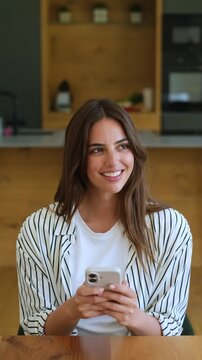Beautiful smiling woman using her smartphone, texting and browsing social media while sitting at a table in her modern kitchen. A positive digital communication and technology concept