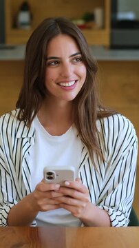 Charming young woman sitting at a table, smiling while using her smartphone for social media, she looks away thoughtfully while communicating online with friends in a modern home