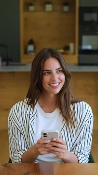 Beautiful smiling woman using her smartphone, texting and browsing social media while sitting at a table in her modern kitchen. A positive digital communication and technology concept
