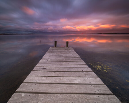 View of a wooden jetty extending into the calm waters of Wilson Inlet at sunset with a dramatic purple and orange cloudy sky in Nullaki, Western Australia, Australia.