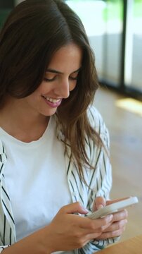 Charming young woman with long brown hair smiling while scrolling and typing on her smartphone, enjoying social media communication or browsing the internet in a brightly lit room