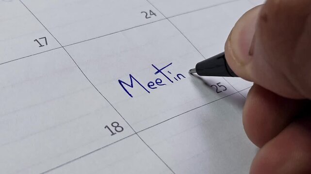 Close up of hand writing the word 'Meeting' with a blue pen on a paper calendar