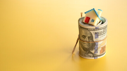 An upright rolled wad of United States one hundred dollar bills with a miniature house toy nestled inside on a solid yellow surface. © MohamadFaizal