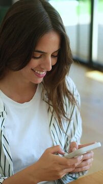 Beautiful young woman smiling while texting on her smartphone, enjoying a pleasant conversation online, sitting at a wooden table in a cafe, absorbed in her digital communication