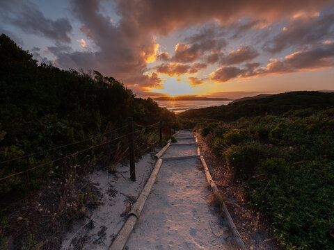 View of a sandy path leading towards the ocean at sunset with lush coastal vegetation and a dramatic orange sky in Nullaki, Western Australia, Australia.