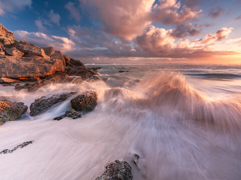 View of Nullaki coastline with waves crashing against rocky outcrops on white sand under a dramatic sunset sky, Nullaki, Western Australia, Australia.