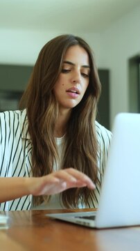 Young professional woman concentrating while typing on her laptop computer, sitting at a wooden desk in a modern home office setting, focused on completing her digital project