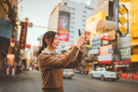 Side view of happy Asian woman smiling and holding smartphone to take photos at Chinatown Bangkok, beautiful young female traveler taking picture of street food and city life during evening holiday