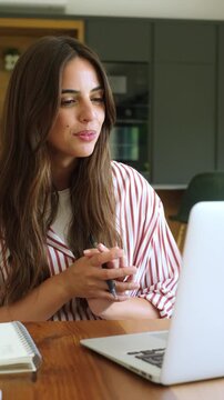 Smiling young businesswoman participating in a video conference call on her laptop, actively discussing and gesturing while working remotely from her modern home kitchen office space