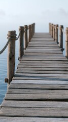 Fototapeta premium Wooden pier extending over calm water with fog in the background, featuring weathered planks and rope railings, creating a tranquil and serene atmosphere