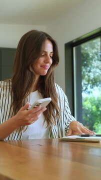 Beautiful young woman sitting at a wooden table in her modern home. Smiling as she types and scrolls on her smartphone. Managing her schedule with a notebook nearby. Enjoying a remote work lifestyle