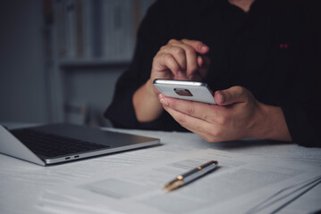 Fototapeta premium Close up of businessman hands using smartphone late at night. Unrecognizable person texting or browsing internet at dark office desk with laptop, document, and pen. Concept of overtime work.