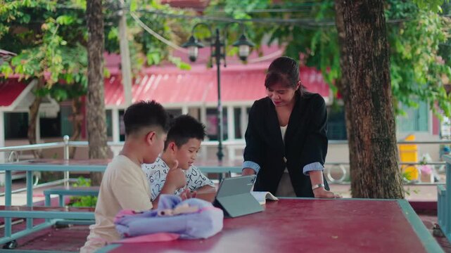Smart Asian primary students studying and reviewing lessons with a helpful female teacher outside the classroom. Cheerful young boys learning together with school textbooks.