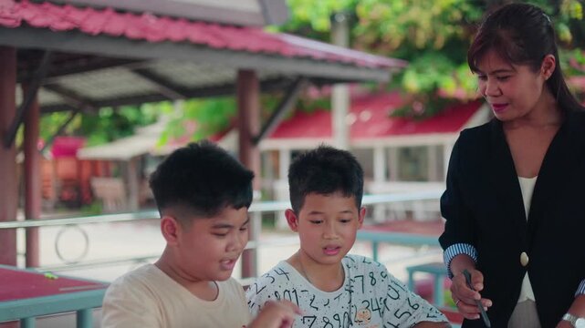 Smart Asian primary students studying and reviewing lessons with a helpful female teacher outside the classroom. Cheerful young boys learning together with school textbooks.