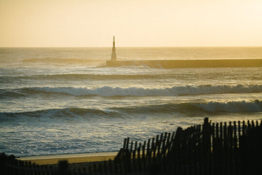 Golden sunset over the Atlantic Ocean with rolling waves, a breakwater lighthouse, and weathered dune fencing framing a windswept coastal shoreline.