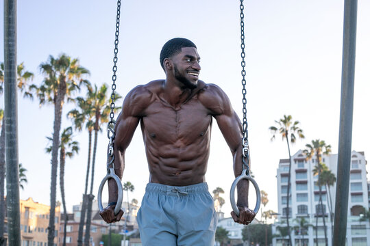 Man exercising on rings outdoors, showcasing muscular physique.