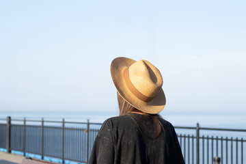 Woman wearing a straw hat stands by a railing overlooking the ocean, with a clear blue sky and calm water visible in the background © Anastasiia