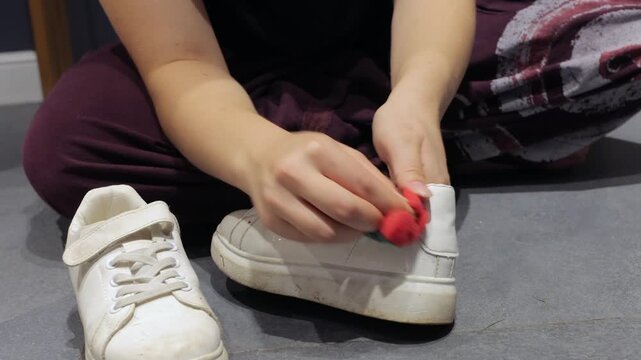 A red sponge wipes grime from a white casual shoe on a gray indoor floor.